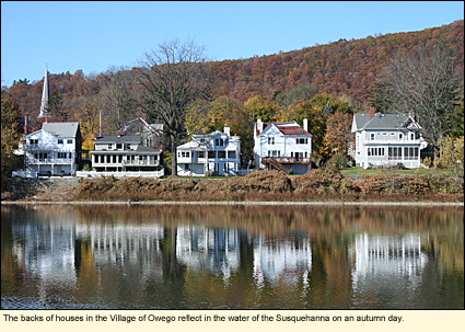 The backs of houses in the Village of Owego, New York reflect in the water of the Susquehanna River on an autumn day.