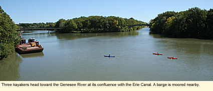 Three kayakers head toward the Genesee River at its confluence with the Erie Canal. A barge is moored nearby.